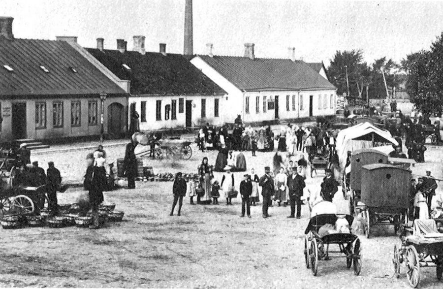 Svartvitt fotografi från slutet av 1800-talet. På torget är många samlade bland vagnar dragna av hästar. I bakgrunden ligger gathus. 