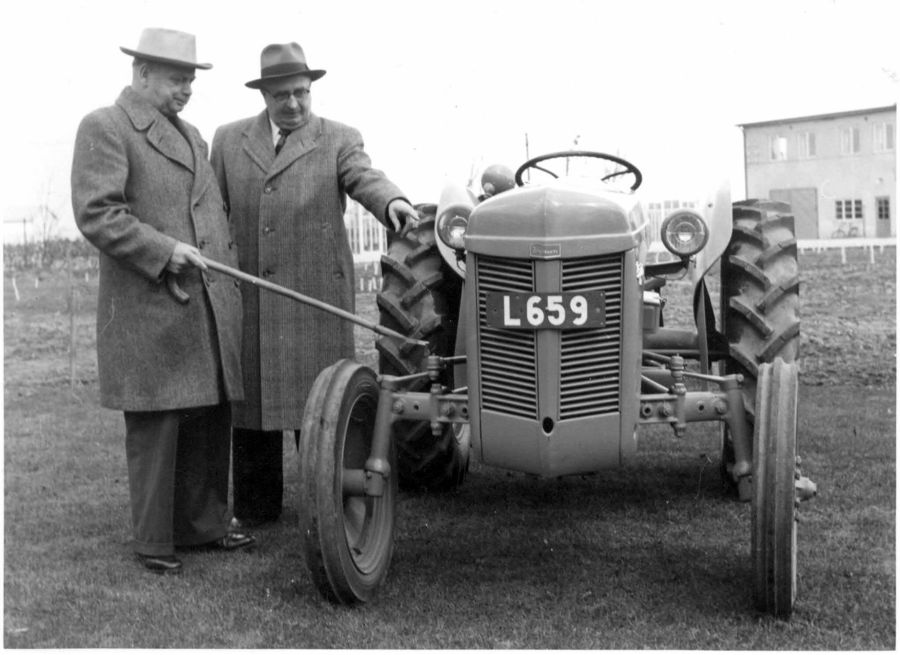 Svartvit bild med två män iklädd hatt och rock inspekterar en gammal traktor av modellen "Grålle"