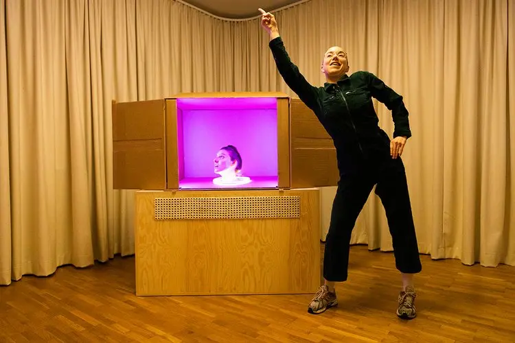 En föreställningen med en kvinna i svart overall som dansar sidan om en låda upplyst i lila ljus med ett kvinnohuvud som sticker upp.